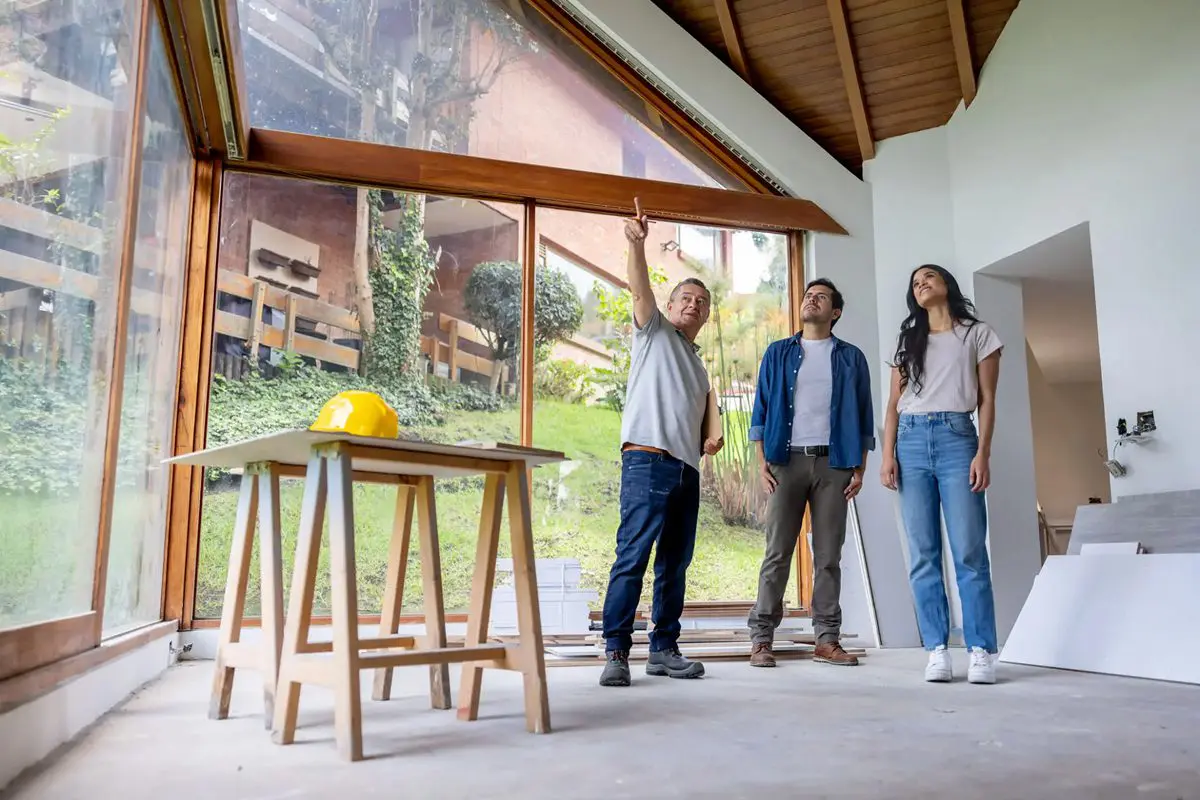 Three people discussing in a modern, open room with wooden stools and large windows.