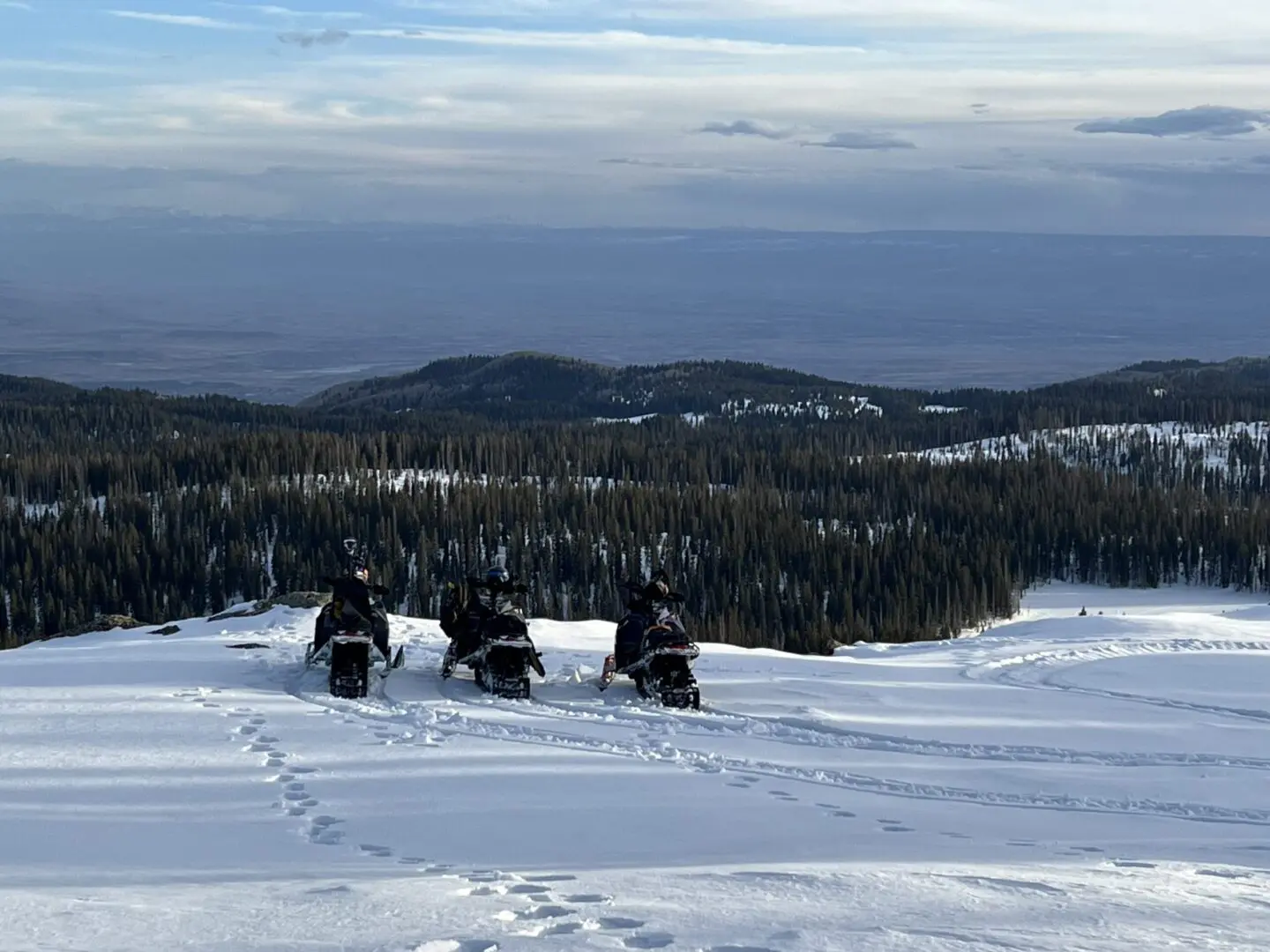 Three snowmobilers traversing snowy hills under a cloudy sky.
