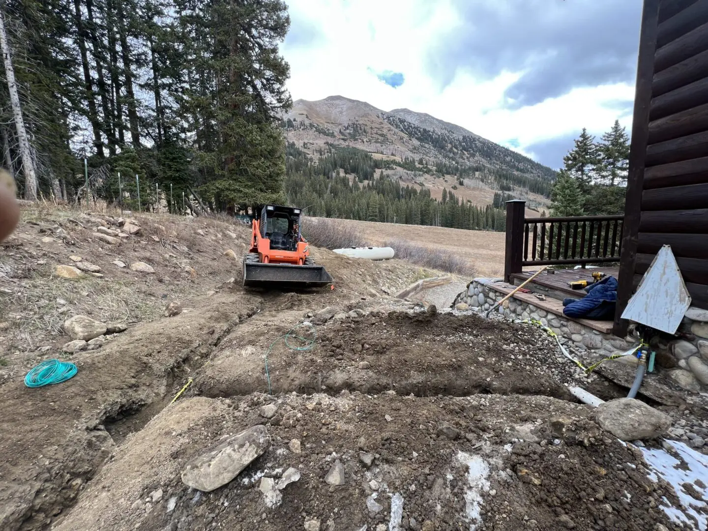 Excavator working on a rocky terrain near a wooden fence and mountainous backdrop.